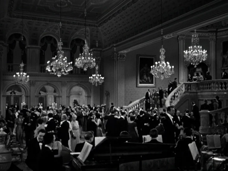 Saboteur (1942) movie still: establishing — A grand ballroom features a curved staircase and multiple large crystal chandeliers hanging above. A…
