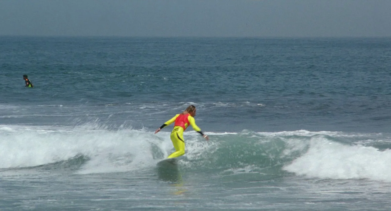 A Scene at the Sea (1991) movie still: wide — A surfer rides a breaking wave in the middle of a large blue ocean. Another distant surfer floats in…