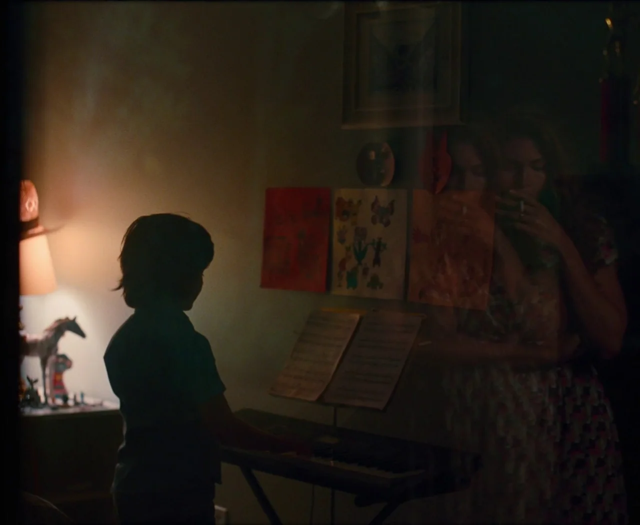 Monica (2022) movie still: medium — A dimly lit room contains a young boy playing a digital keyboard near a desk. Children's drawings are taped to…