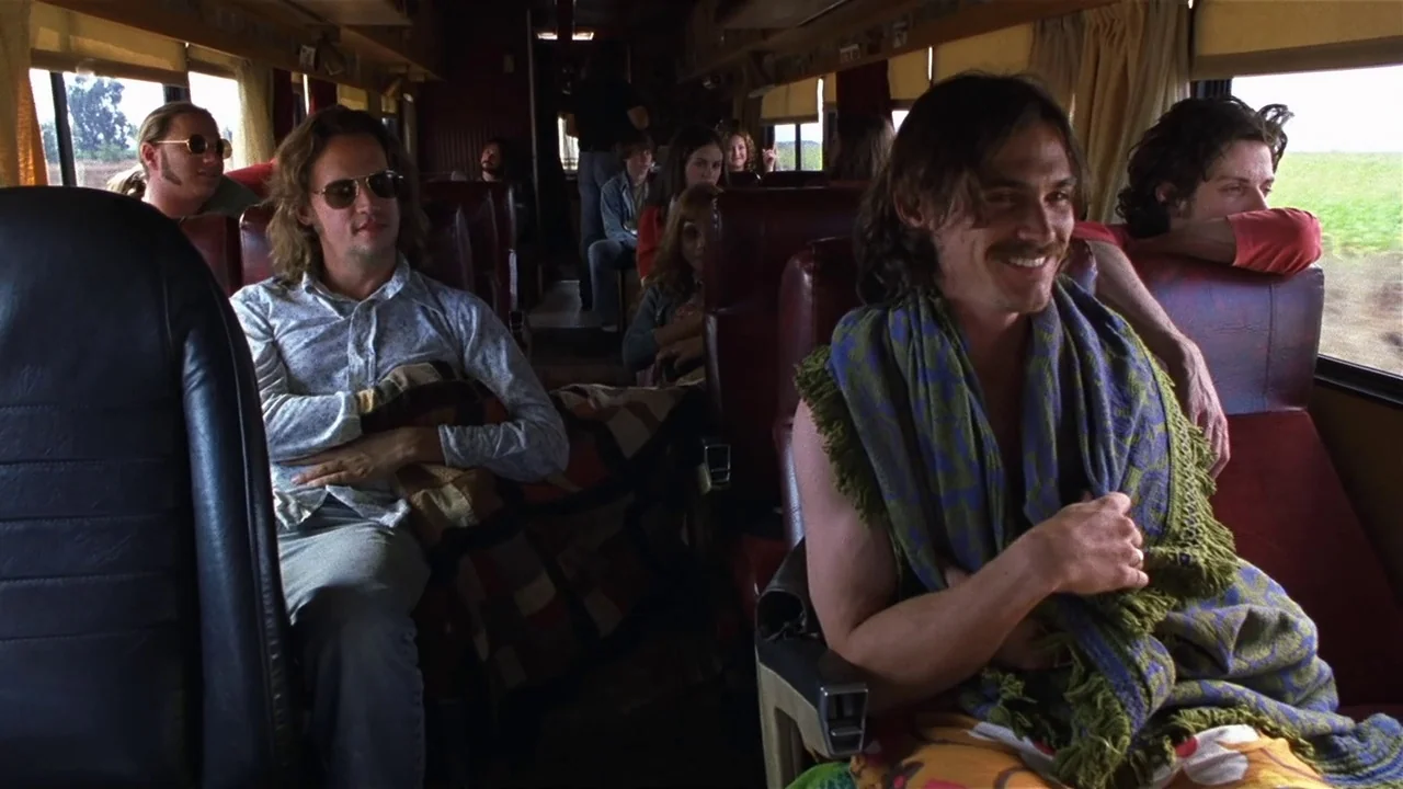 Almost Famous (2000) movie still: medium — An interior shot of a vintage train carriage filled with passengers sitting on red seats. Several young…