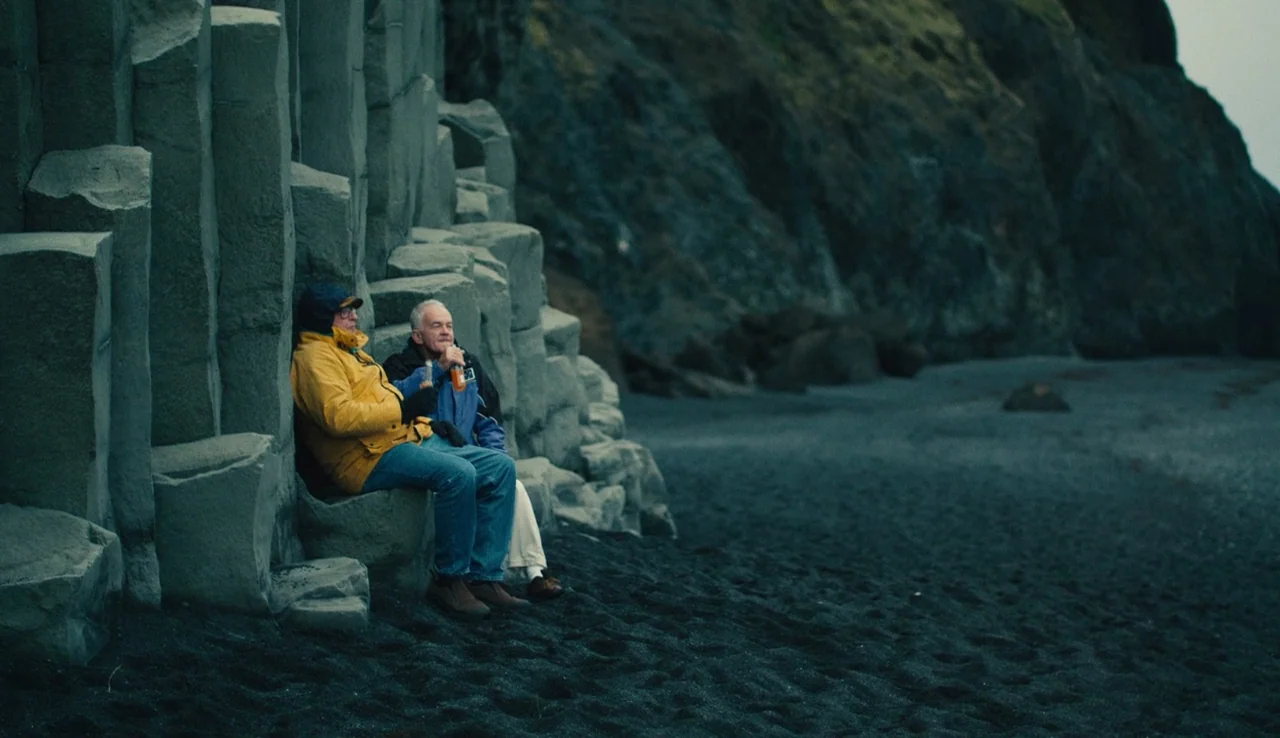 Land Ho! (2014) movie still: fullbody — Two men sit against basalt columns on a dark volcanic sand beach. They hold small bottles while sitting on…