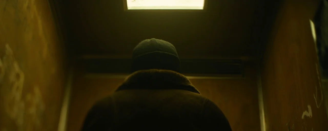 Petrov’s Flu (2021) movie still: medium — A cramped, dimly lit metal box shows graffiti on the side panels. A lone figure stands facing the closed…
