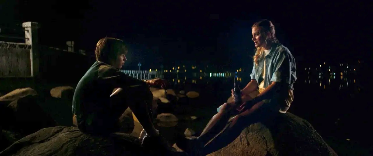 Summer of 84 (2018) movie still: fullbody — A dark, rocky shoreline at night overlooks a body of water with distant lights. A young man and a woman…