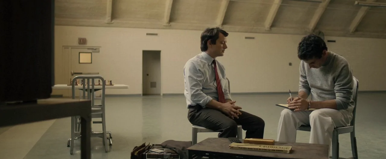 No Man of God (2021) movie still: medium — A sparse, large room with high vaulted ceilings and white painted concrete block walls. Two men sit on…