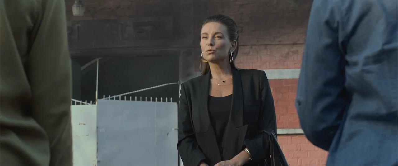 Mirreyes contra Godínez 2: El retiro (2022) movie still: medium — A woman stands before a weathered red brick wall near a metal gate. She faces an…