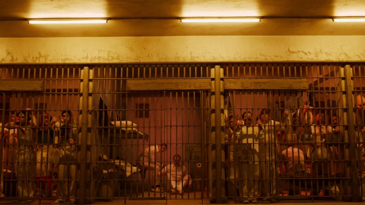 The Laundromat (2019) movie still: wide — A dense prison block features rows of metal bars and numbered cell compartments. Men in matching white…