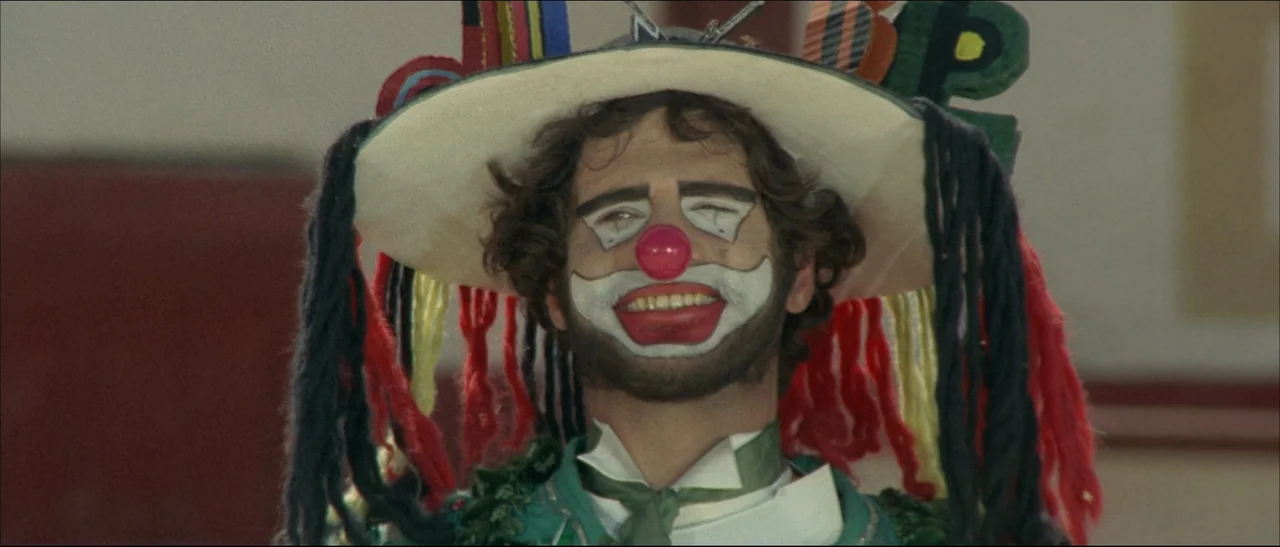 The Mercenary (1968) movie still: closeup — A person in detailed clown makeup and a decorated hat stands outdoors. The subject looks directly…