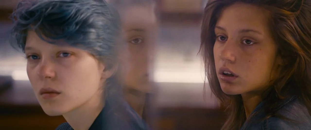 Blue Is The Warmest Colour (2013) movie still: closeup — Two women sit close together in a softly lit interior space. Their faces overlap in the…
