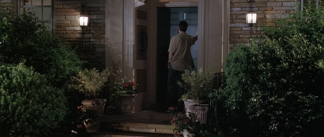 A Walk to Remember (2002) movie still: medium — A stone-front residential entrance at night features potted plants flanking a central door. A man…
