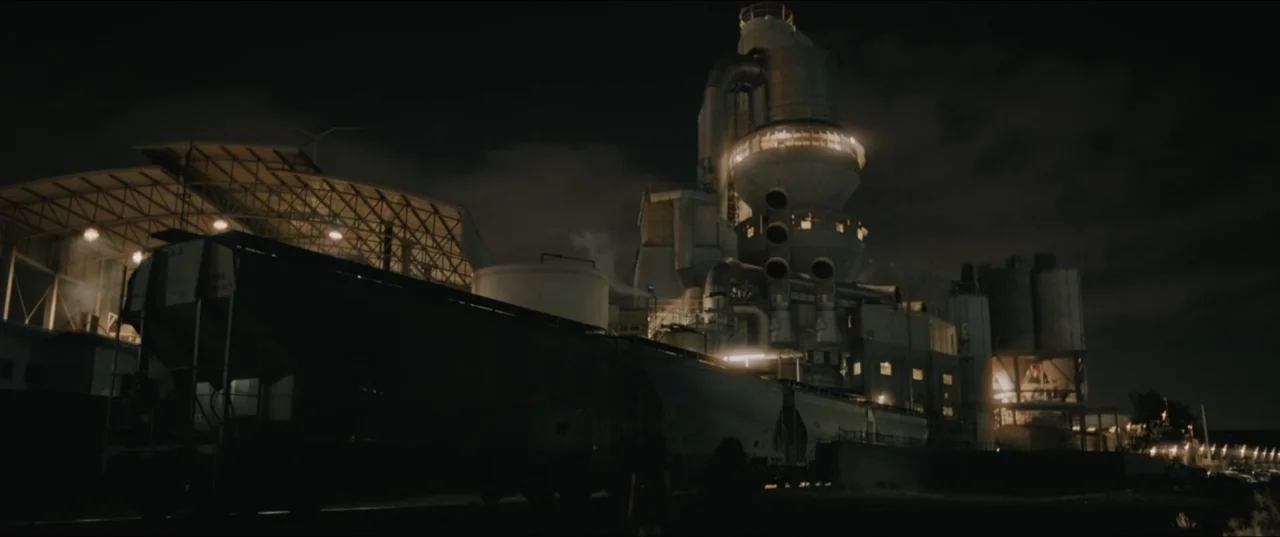 Death to Summer (2021) movie still: establishing — A large industrial facility with tall towers and pipes is situated at night. A freight train car…