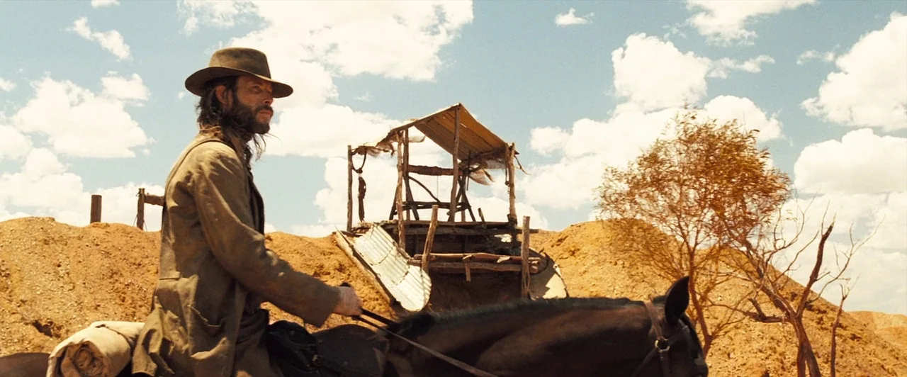 The Proposition (2005) movie still: medium — A man rides a horse through a barren desert landscape with orange, hilly terrain. A small, derelict…