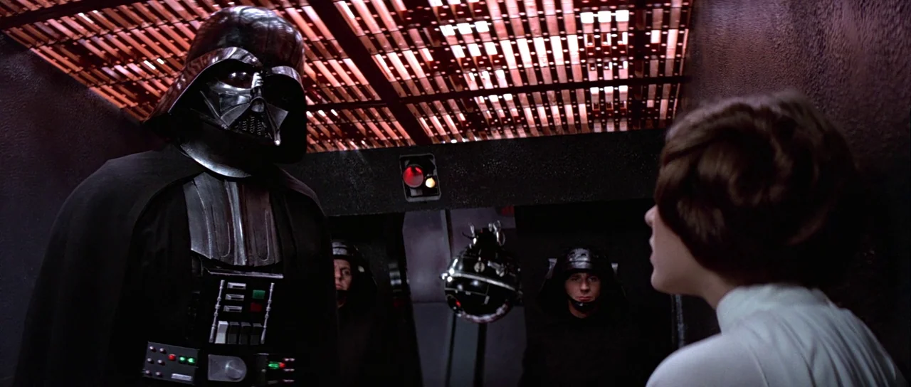 Star Wars (1977) movie still: medium — A dimly lit metallic corridor features a red overhead lighting grid and dark walls. A tall black armored…