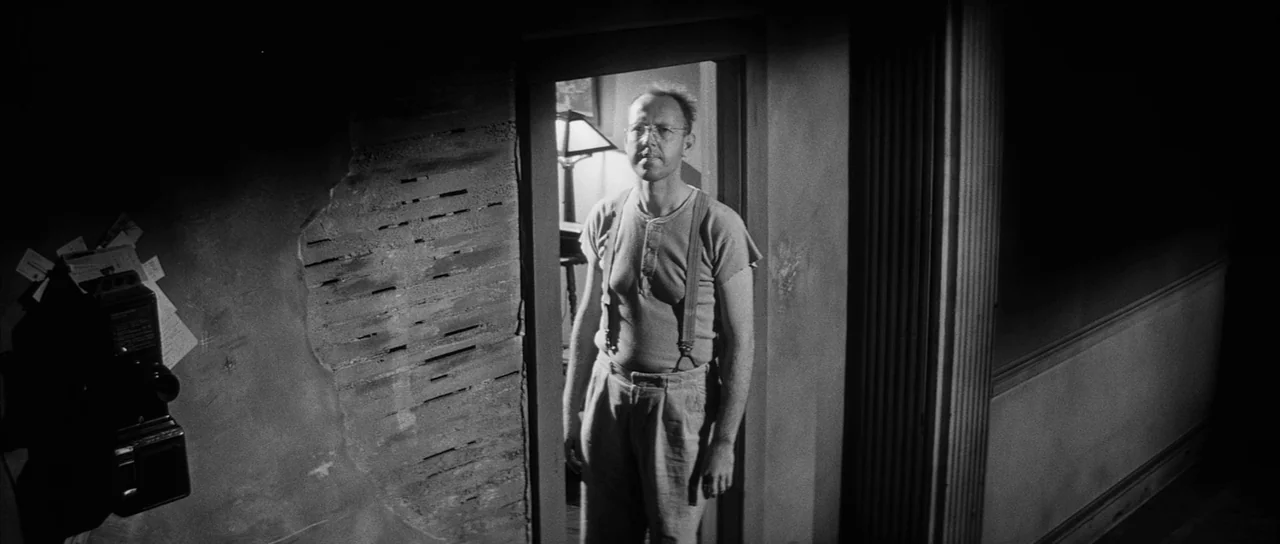 The Tarnished Angels (1957) movie still: medium — A dimly lit narrow hallway shows a wall with missing plaster exposing wooden laths. A man in a…