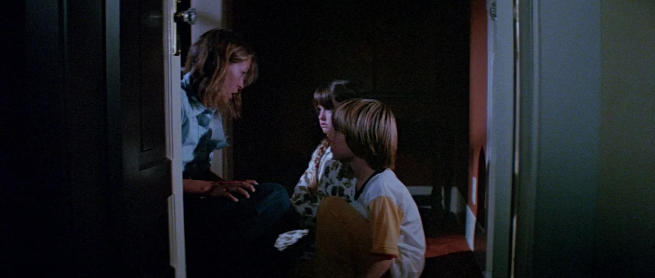 Halloween II (1981) movie still: medium — A dimly lit residential hallway at night features a woman kneeling before two children. She addresses the…