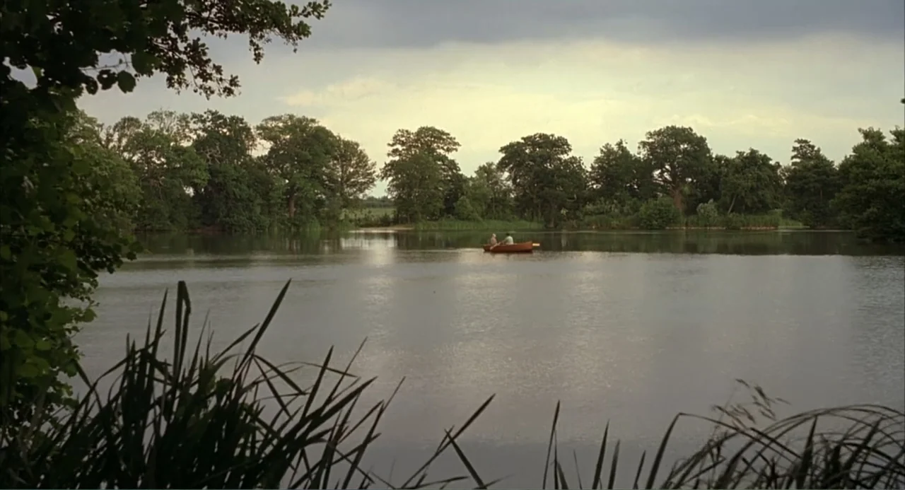 Scoop (2024) movie still: wide — A wide, still lake is framed by tall green deciduous trees under an overcast sky. Two small figures occupy a wooden…