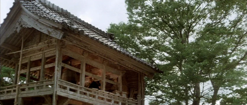 The Horrors of Malformed Men (1969) movie still: wide — A wooden Japanese belfry with traditional tile roof stands beside a large green tree. The…