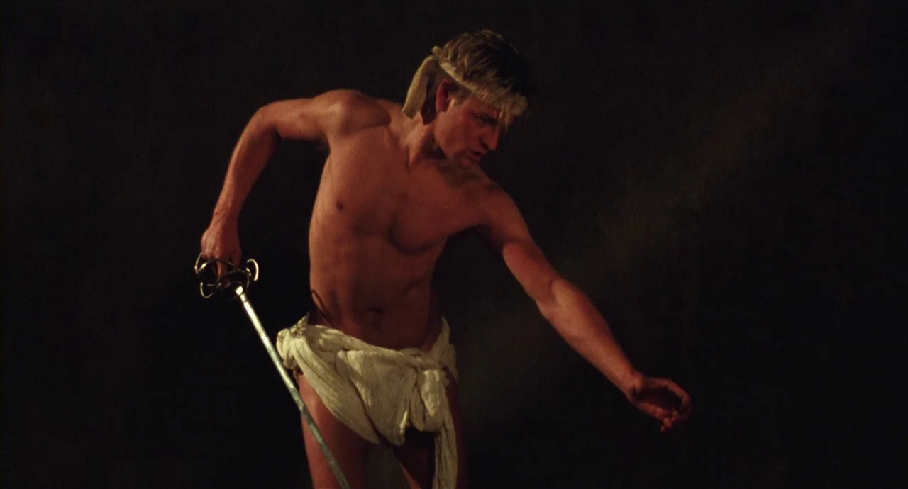 Caravaggio (1986) movie still: medium — A man stands before a featureless dark backdrop with no visible floor or ceiling. The shirtless man wears a…
