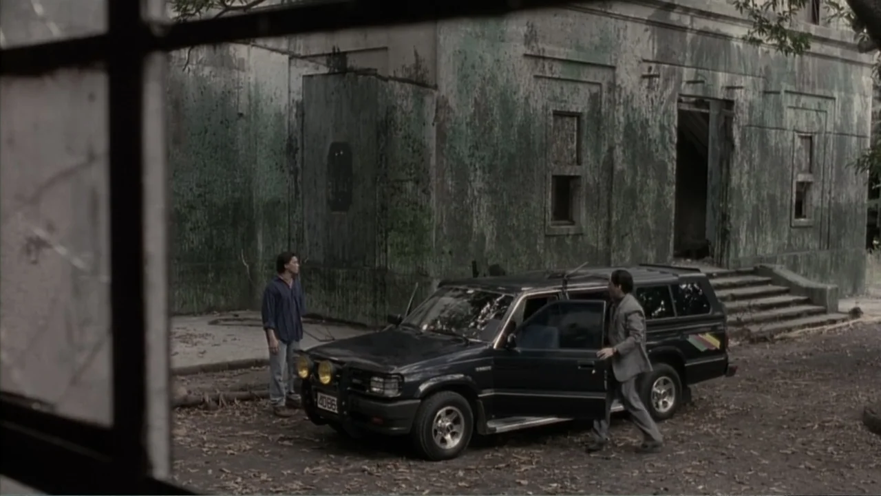 Behind Enemy Lines (2001) movie still: wide — A dark SUV is parked in front of a crumbling, moss-covered concrete building. Two men interact near…