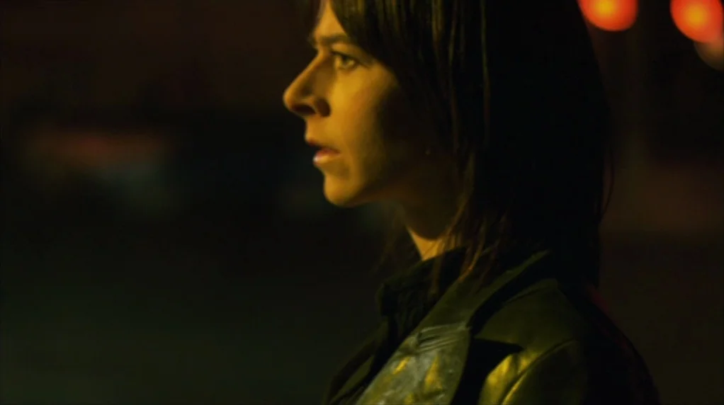 Red Road (2006) movie still: closeup — A dark outdoor environment at night lit by a single warm external light source. A young woman stands looking…