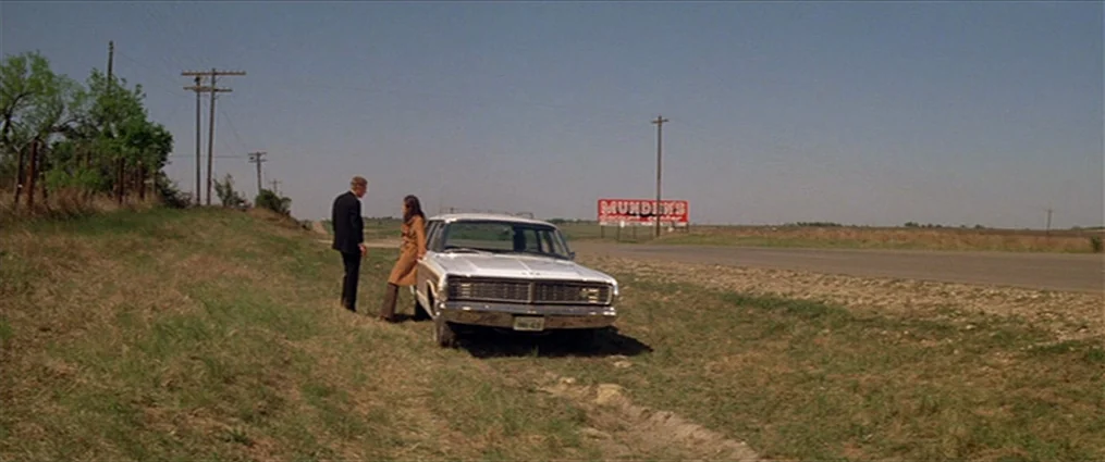 The Getaway (1972) movie still: wide — An arid roadside area with dry grass sits beneath a clear blue sky. A man and a woman stand together by a…