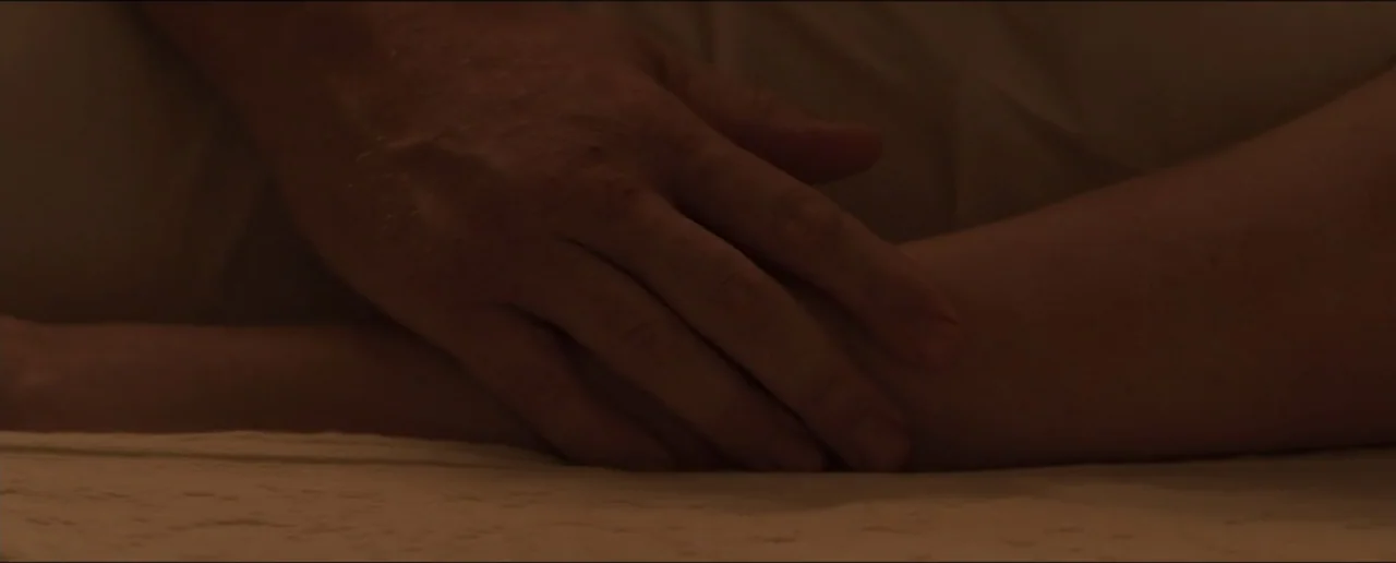 Hostages (2017) movie still: closeup — A dark, out-of-focus indoor space features beige bedding material. A person's hand rests calmly upon a forearm.