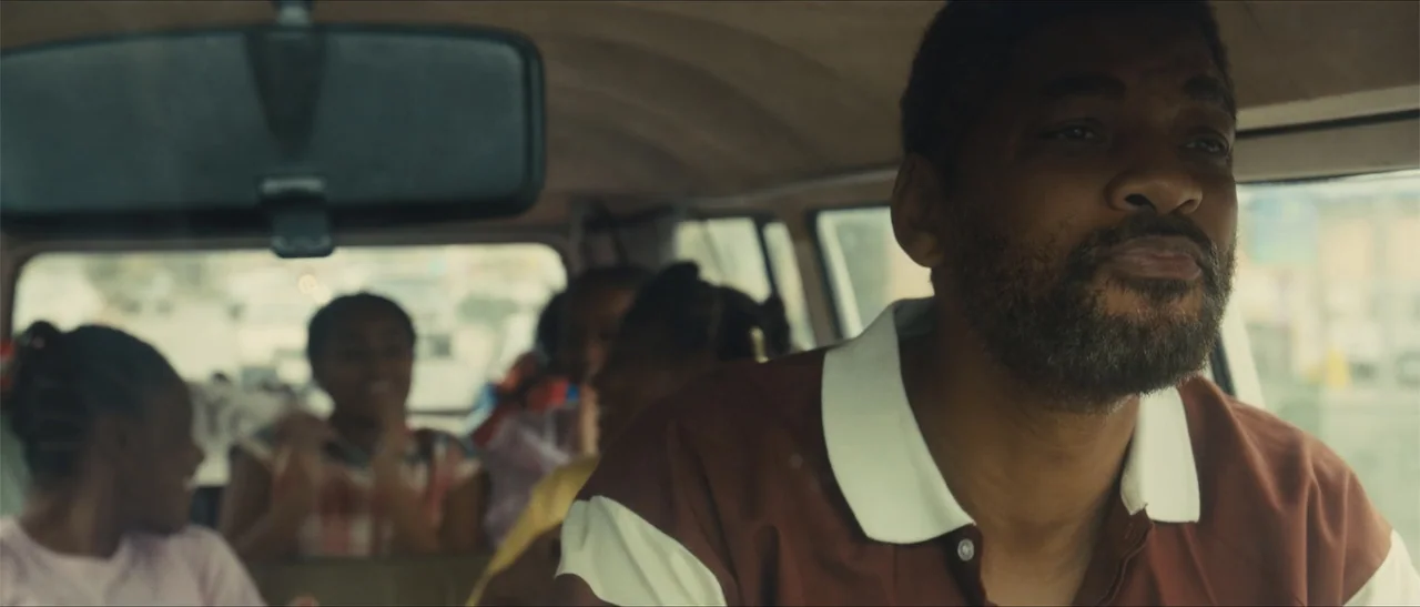 King Richard (2021) movie still: medium — Inside a moving vehicle, a man sits in the driver seat looking ahead. Multiple children are visible in the…