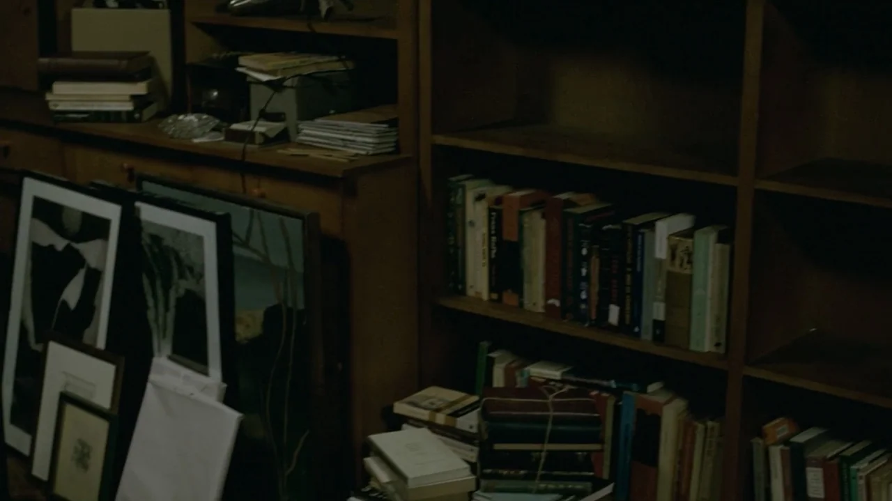 I'm Still Here (2024) movie still: wide — A wooden bookcase filled with numerous books and framed art prints stacked on floor. The library shelves…
