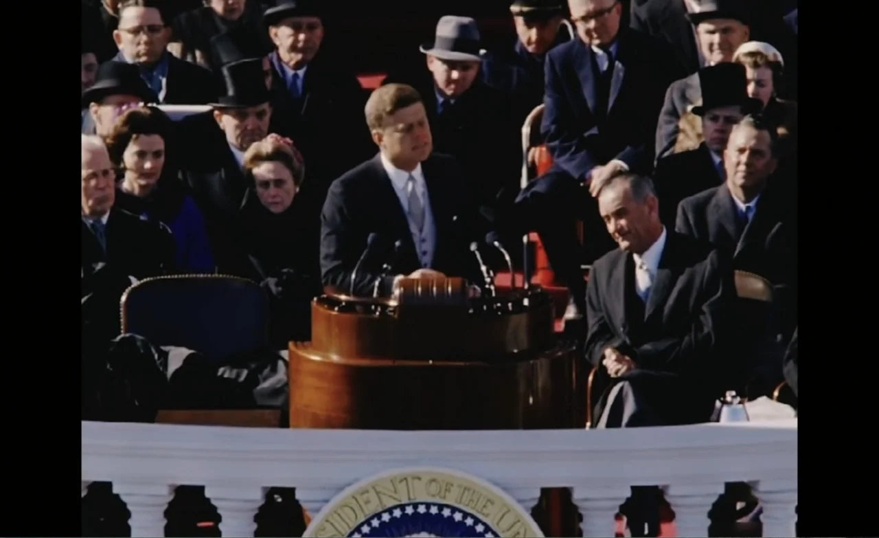 Let the Canary Sing (2023) movie still: medium — A man speaks from behind a podium with a presidential seal in a crowded area. He wears a formal…