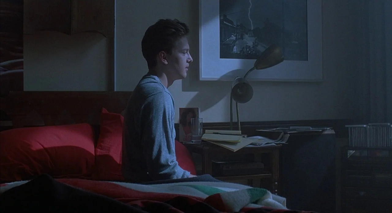 Less Than Zero (1987) movie still: medium — A dimly lit room at night features a bed with red pillows. A man sits on the side of the bed next to a…