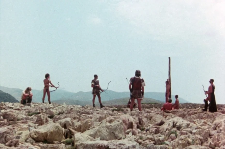 Sebastiane (1976) movie still: wide — A rocky hillside under a pale, overcast sky serves as a sparse, desolate setting. Several men in primitive…