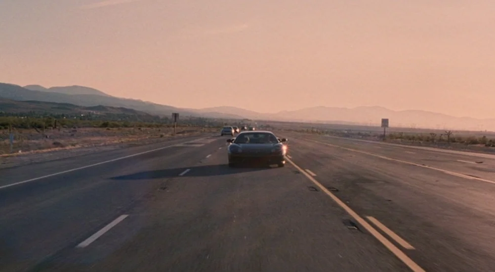Somewhere (2010) movie still: wide — A vast, multi-lane desert highway extends toward distant mountains under a warm, late-afternoon sky. A black…