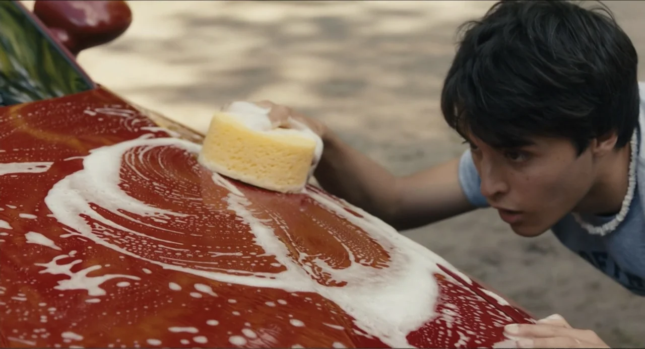Hell of a Summer (2025) movie still: medium — A sunlit outdoor area features a red vehicle hood covered in white soapy foam. A young man with dark…