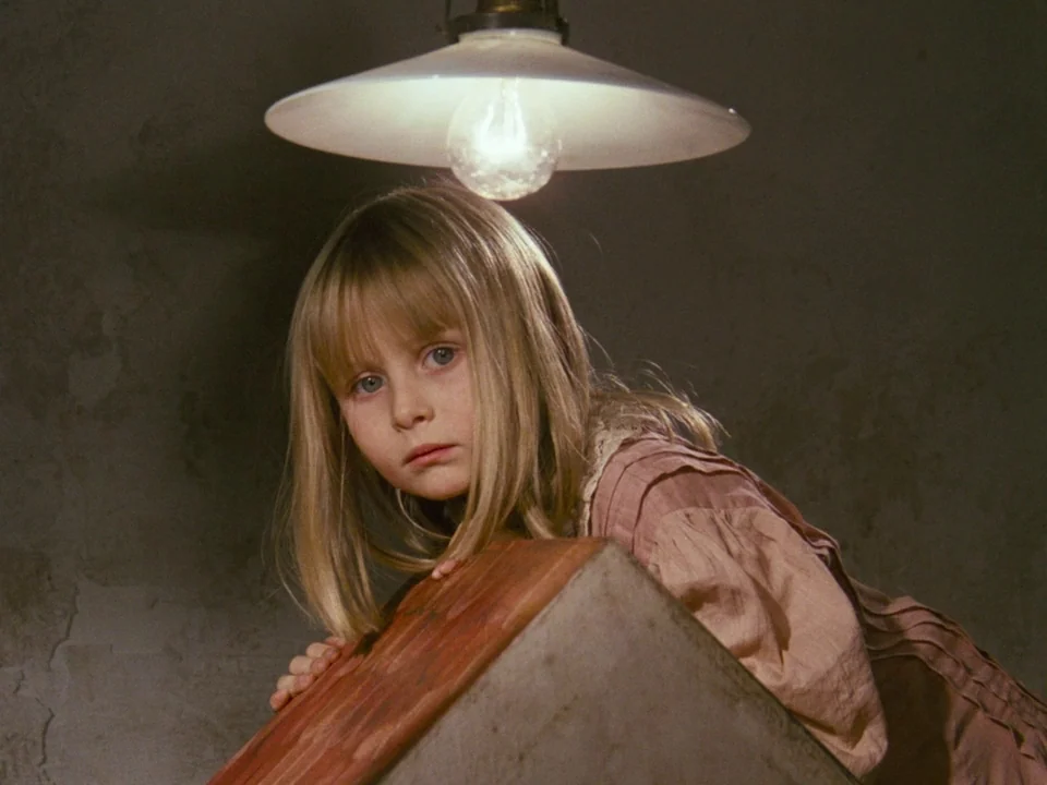 Alice (1988) movie still: medium — A dimly lit, bare room features a rough grey wall and a concrete structure. A young girl with blonde hair looks…