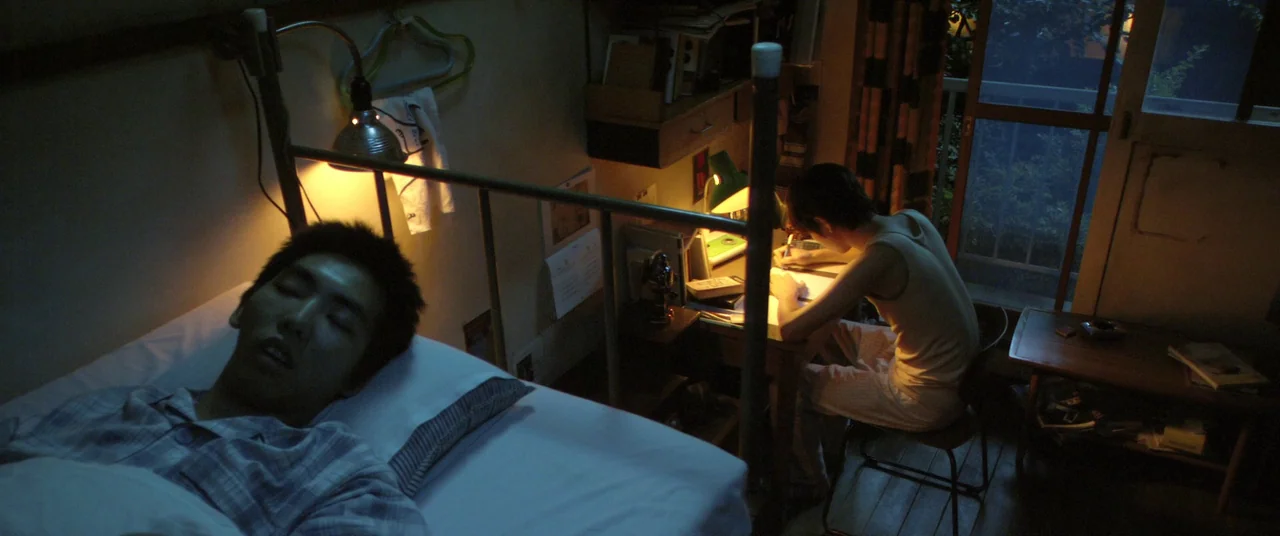 Norwegian Wood (2010) movie still: medium — A cramped, dimly lit room contains a metal-frame bunk bed and a small desk. One person lies sleeping on…