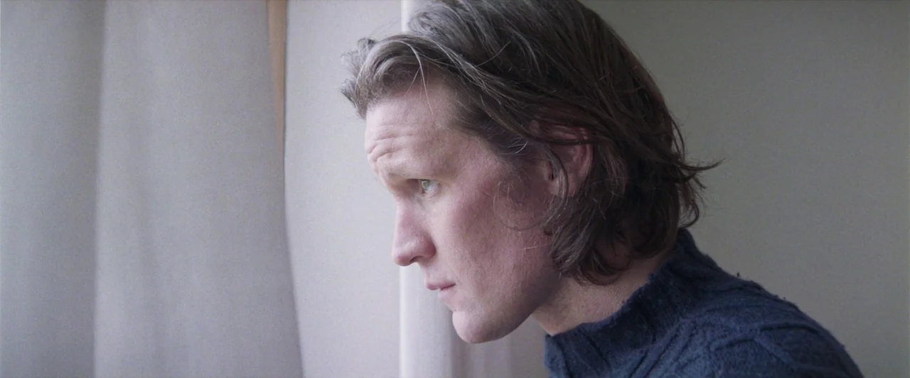 Starve Acre (2023) movie still: closeup — A light-filled room with plain wall surfaces and a sheer curtain. A man in a blue sweater turns his head…