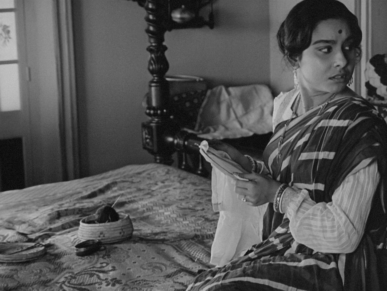 Charulata (1964) movie still: medium — A woman sits on a bed covered by a decorative patterned quilt. She holds a piece of white cloth while looking…