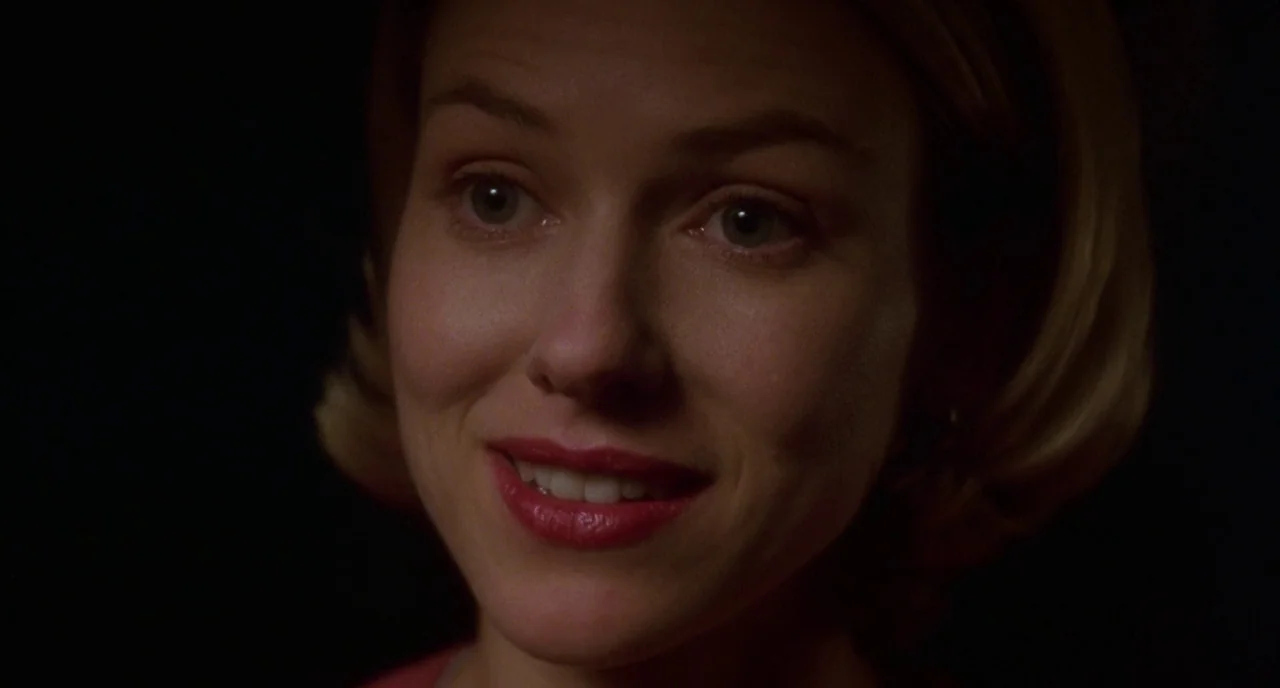 Mulholland Drive (2001) movie still: closeup — A woman stands in a dark room with a solid black backdrop. She smiles directly at the viewer.