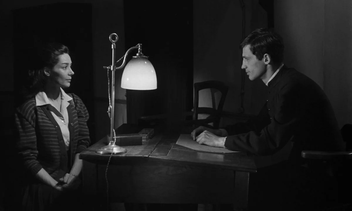 Léon Morin, Priest (1961) movie still: medium — A spare, dimly lit room contains only a wooden table and two simple chairs. A woman and a man sit…