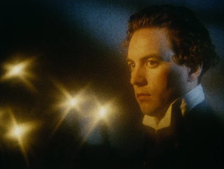 Careful (1992) movie still: closeup — A man stands in a dark room with blurred golden lights in the foreground. He looks to the left while wearing a…