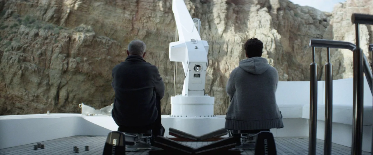 Chevalier (2015) movie still: fullbody — A white boat deck is situated against a tall, jagged beige rock cliff face. Two men sit on the deck facing…