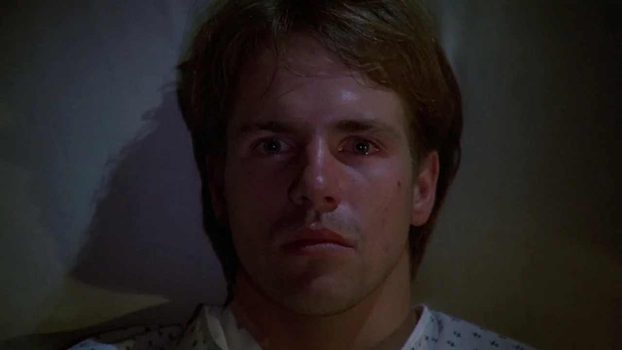 Friday the 13th: A New Beginning (1985) movie still: closeup — A dark hospital room with a plain wall visible behind the subject. A man in a…