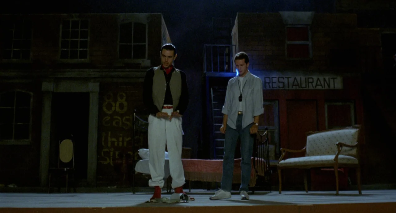 StageFright (1987) movie still: fullbody — A theatrical stage set mimics an outdoor street scene with painted brick facades. Two men stand in the…