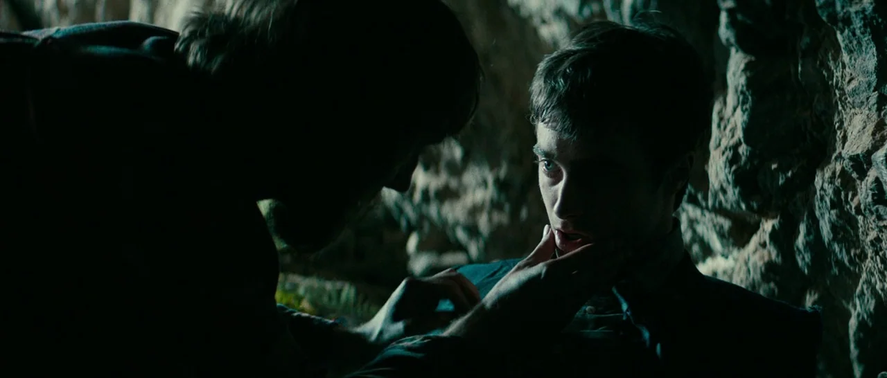 Swiss Army Man (2016) movie still: medium — A dark, cramped stone cave interior with rough, textured rock walls. One man touches the chin of…