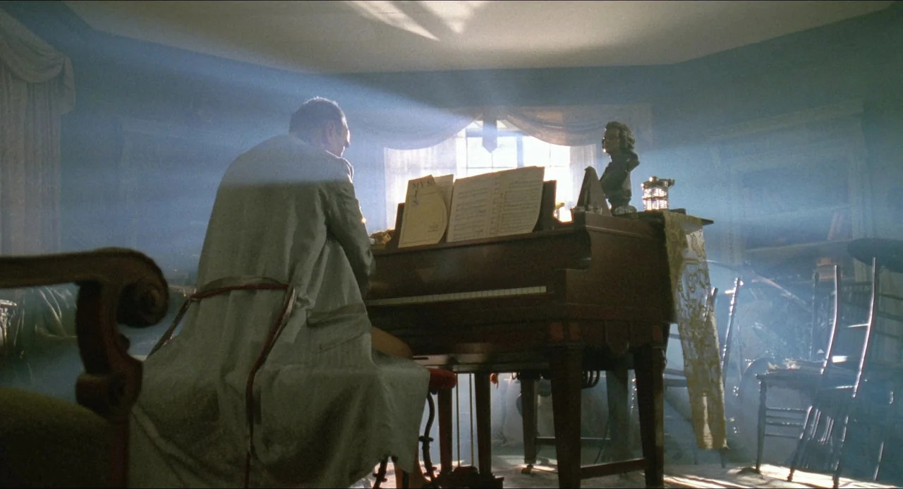 Lolita (1997) movie still: medium — A dust-filled room with hazy light filtering through a central window onto a piano. A man in a robe plays the…