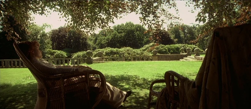 The House of Mirth (2000) movie still: wide — A manicured estate lawn is bordered by a stone balustrade and dense trees. A woman relaxes in a wicker…