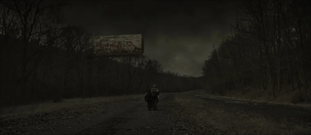 The Road (2009) movie still: wide — An abandoned rural road is surrounded by bare, leafless trees under a dark sky. A man and a child walk away…