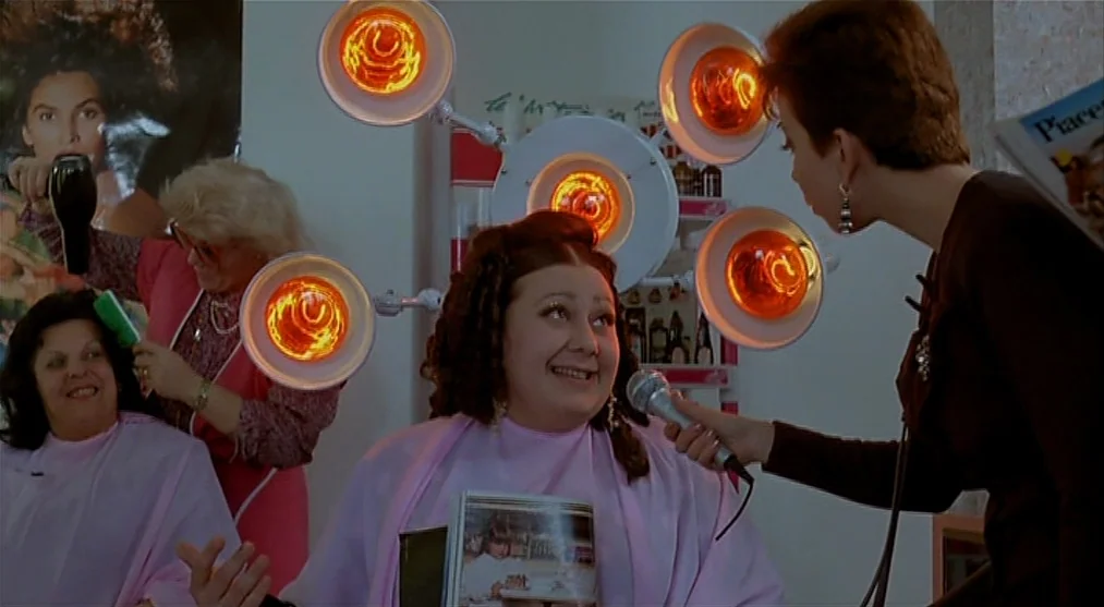 The Voice of the Moon (1990) movie still: medium — A woman with a microphone interviews a customer seated under a multi-lamp salon dryer. The salon…
