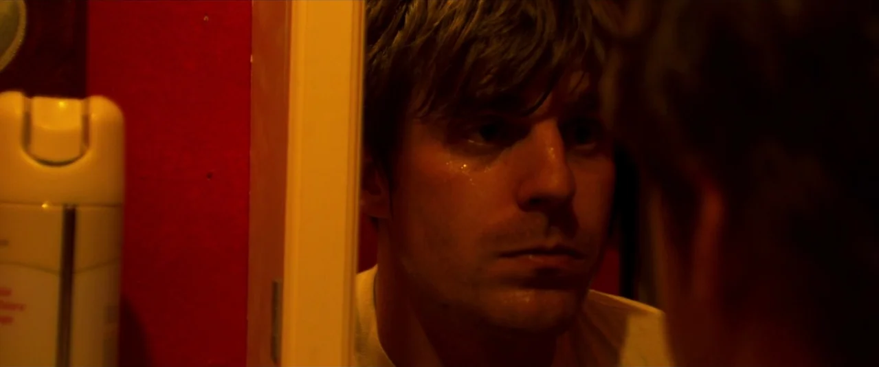 Bellflower (2011) movie still: closeup — A man looks at his reflection in a mirror against a solid red wall. A plastic spray bottle stands in the…