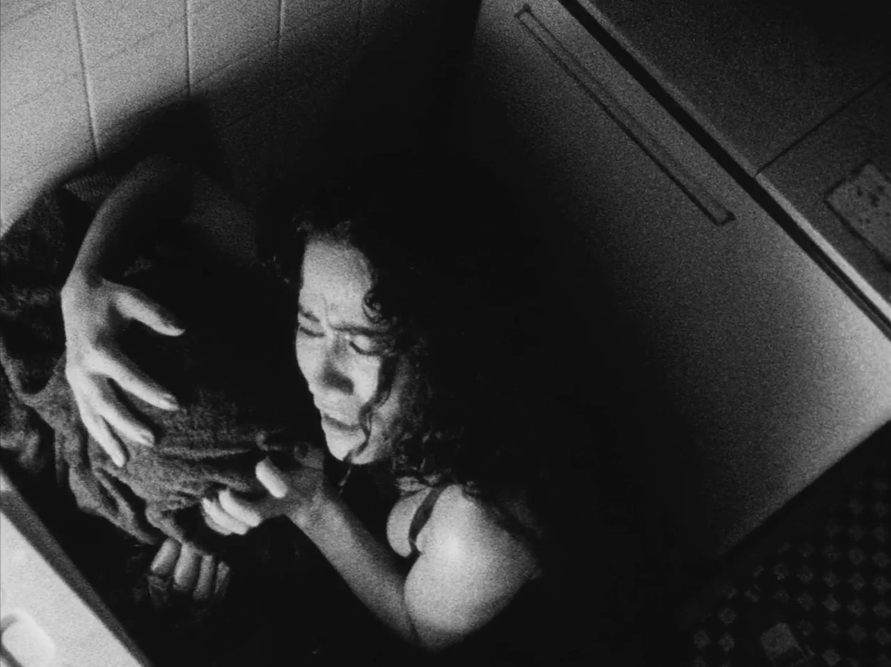 Tetsuo: The Iron Man (1989) movie still: medium — A dark, cramped bathroom with tiled walls and a checkered floor pattern. A woman is huddled on the…