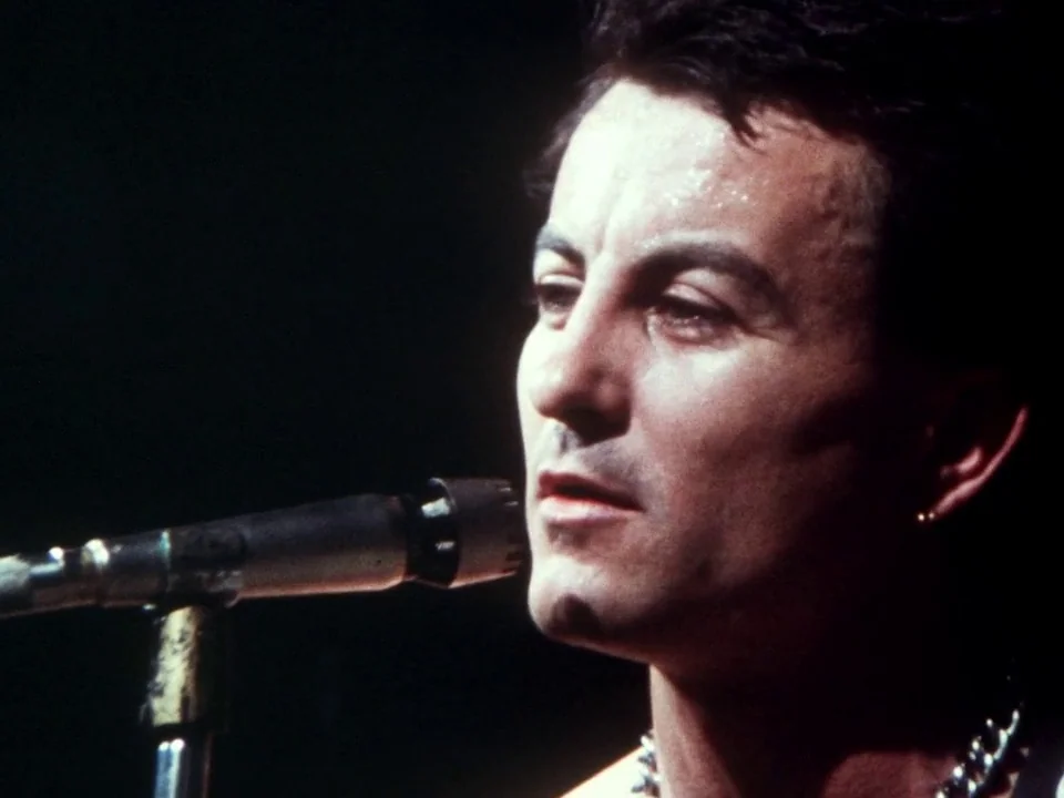 Decline of Western Civilization (1981) movie still: closeup — A musician stands on a dark stage with a microphone in the foreground. He sings while…